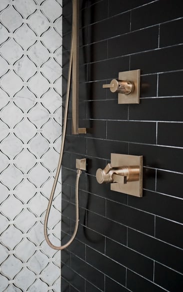 Brushed gold shower fixtures mounted on matte black subway tile and white marble patterned tile in a Herndon, VA bathroom remodel.