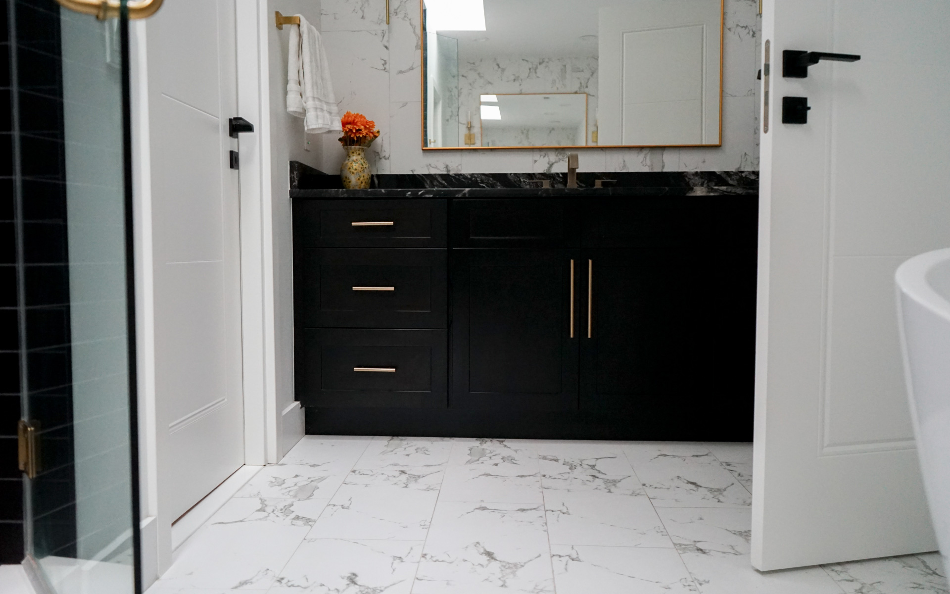 A modern bathroom interior featuring white marble-patterned floor tiles, a sleek black vanity with gold hardware, a large mirror, and white paneled doors.