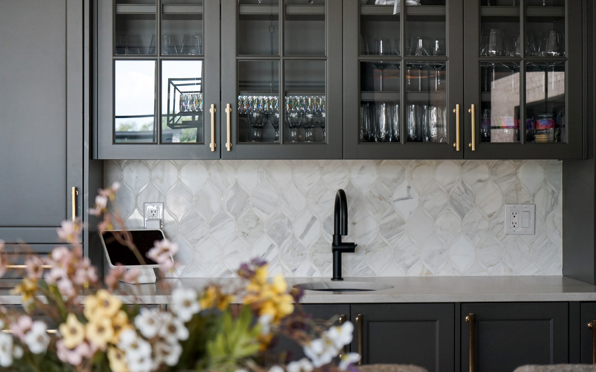 A modern home bar setup with dark grey glass-front upper cabinets, gold handles, a matte black faucet, and a decorative white marble tile backsplash.