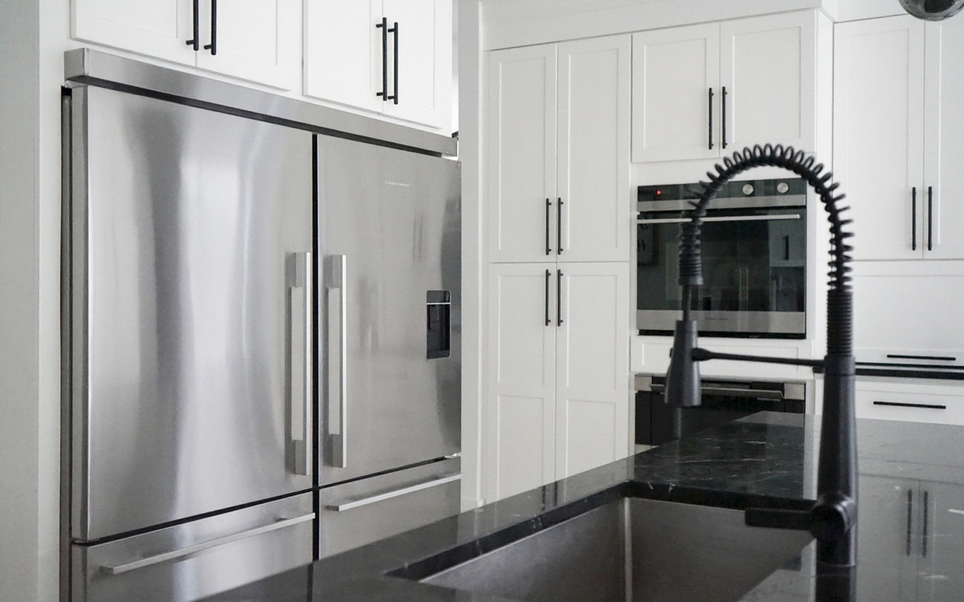 A contemporary kitchen with stainless steel built-in refrigerators, white cabinetry with black handles, built-in ovens, and a black kitchen island with a dark sink and pull-down coiled faucet.