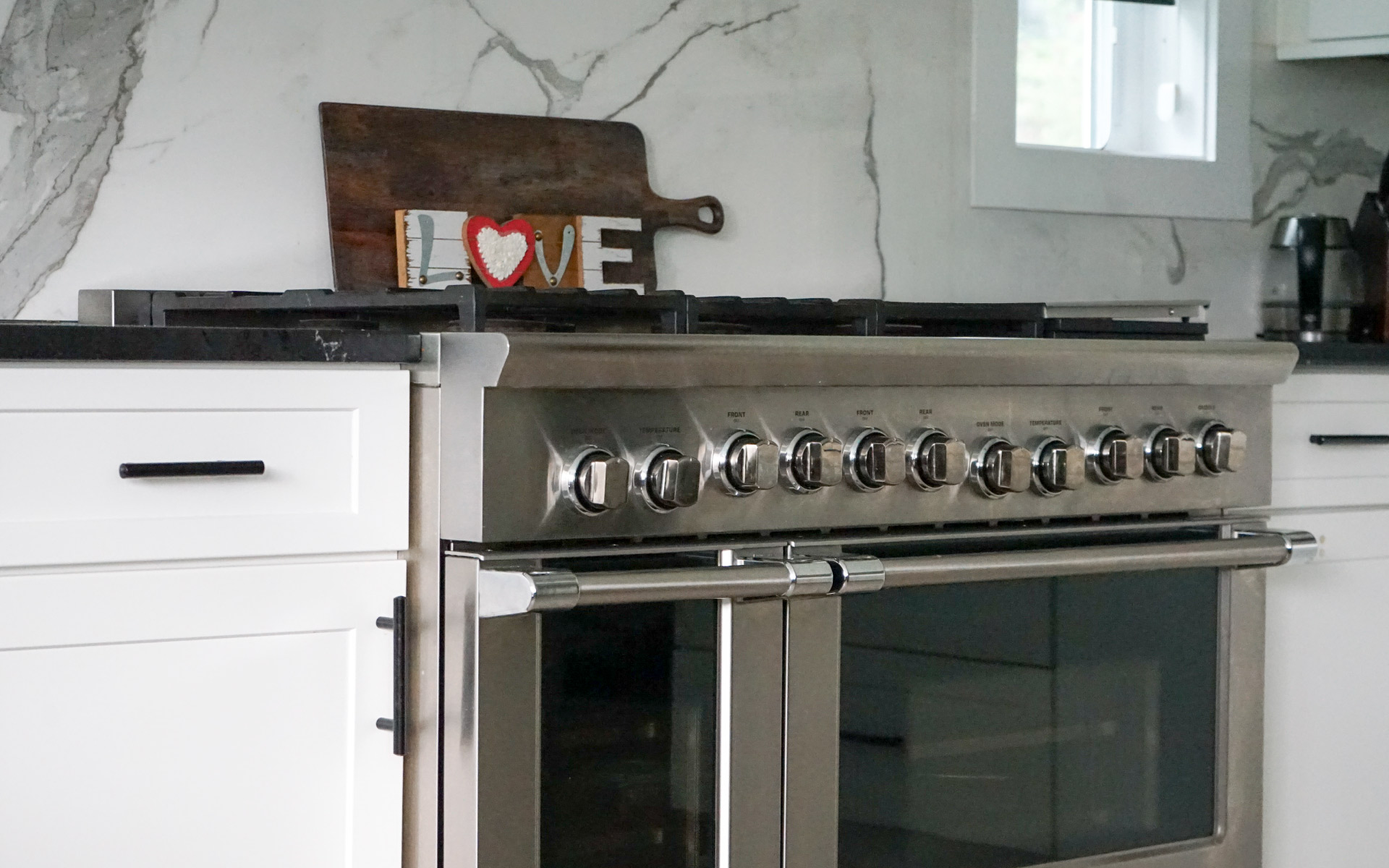 A professional stainless steel gas range with double ovens set against white kitchen cabinets, a marble backsplash, and a wooden cutting board with decorative lettering.