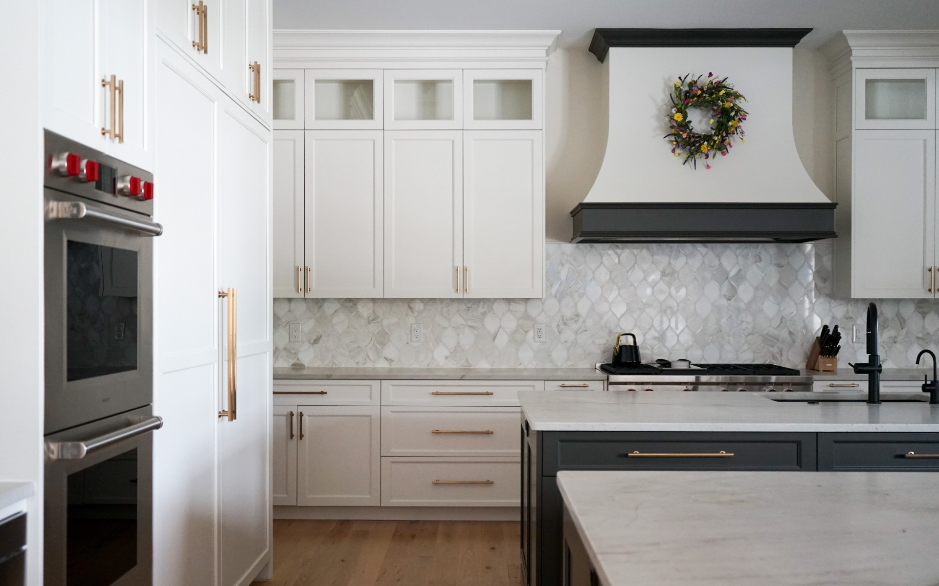 A bright, modern kitchen featuring white cabinets with gold hardware, a dark grey island with a white countertop, stainless steel double wall ovens, and a white range hood with dark trim and a decorative floral wreath.