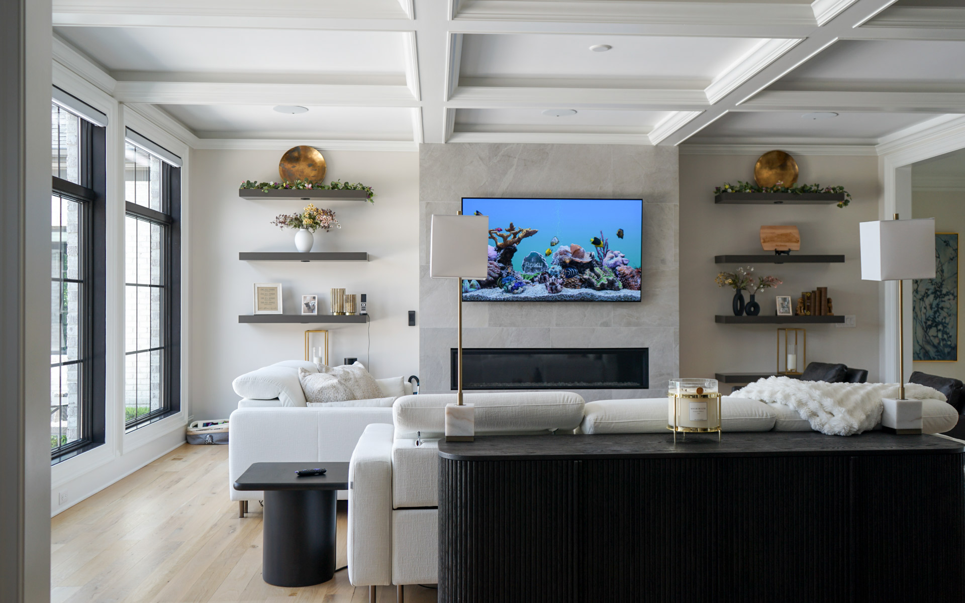 A modern living room with coffer ceiling, a wall-mounted TV displaying an aquarium scene above a linear fireplace, flanked by floating shelves and large windows next to white sectional sofas.