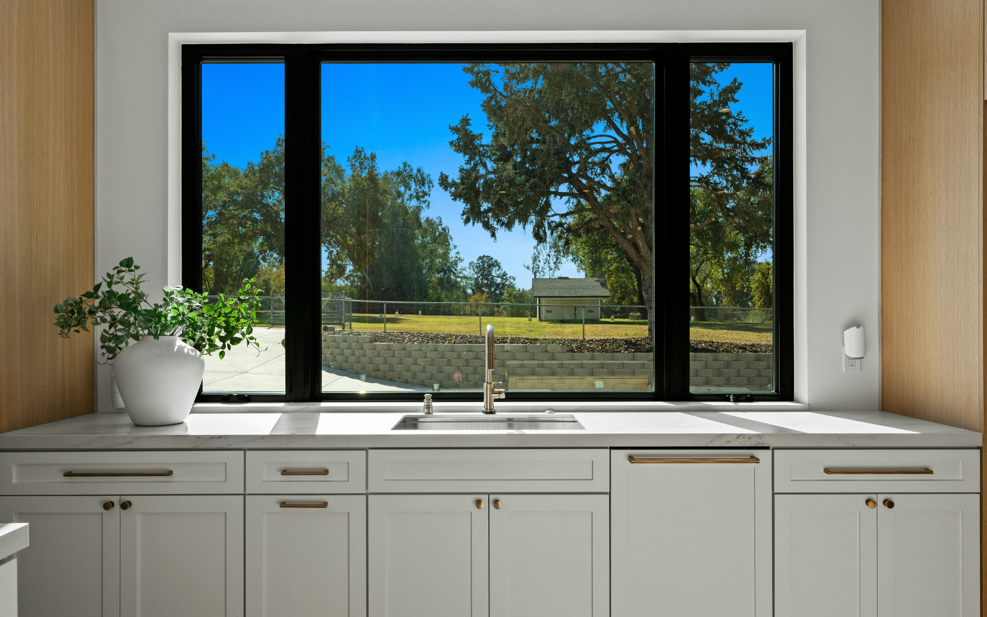 A modern kitchen sink area featuring white cabinetry with gold hardware, a marble countertop with a potted green plant, and a large black-framed picture window overlooking a sunny backyard with green trees.