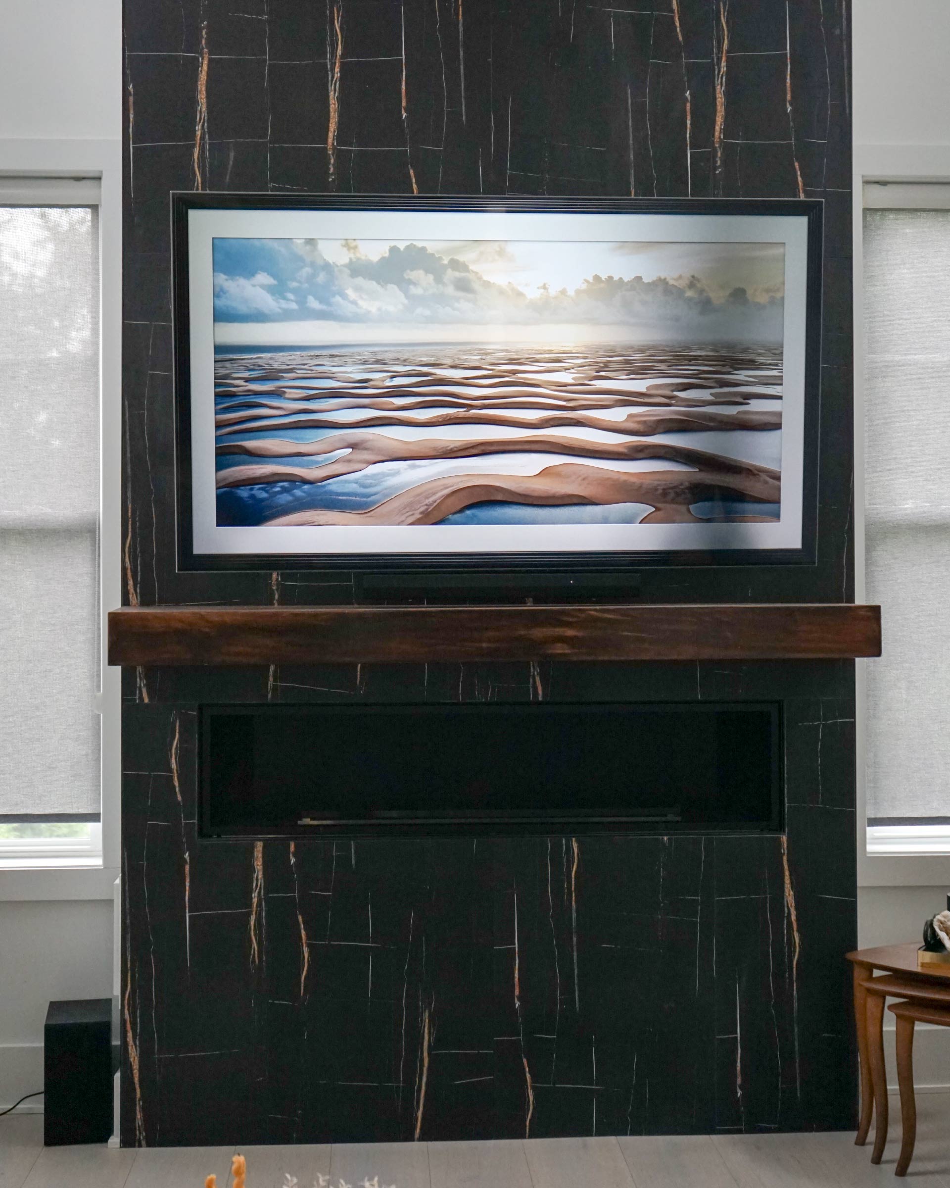A wall-mounted TV displaying a landscape scene above a dark wood mantel and linear fireplace, set against a black vertical tiled accent wall flanked by two large windows.